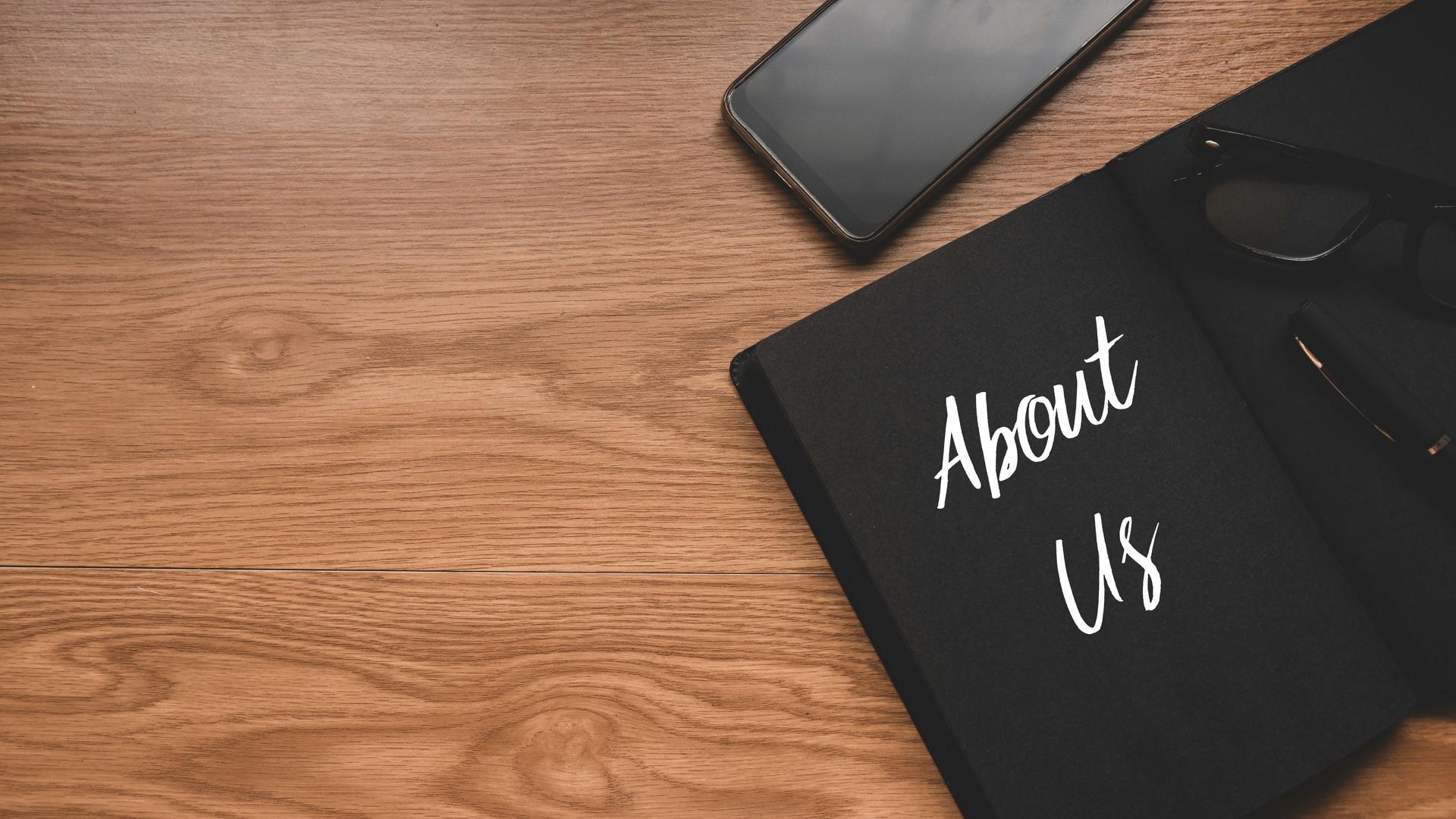 Top view of mobile phone, glasses,pen and notebook written with About Us on wooden background with copy space.